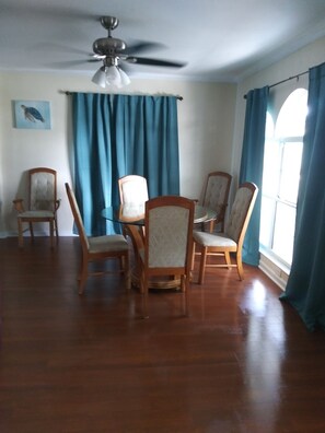 A dining area with a ceiling fan, wooden chairs, a glass table, and blue curtains.