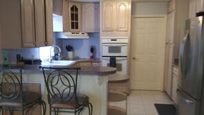 A kitchen with wooden cabinets, a stainless steel refrigerator, a built-in oven, and a sink. There are two bar stools and a small rug on the floor. - Orange Blossom Refuge (Pinellas County)