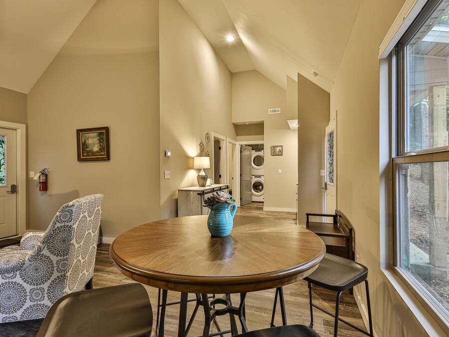 A dining area with a round table, chairs, and a vase. A patterned chair is visible. A door with a stained glass window is on the left. A window with a view of the outside is on the right.