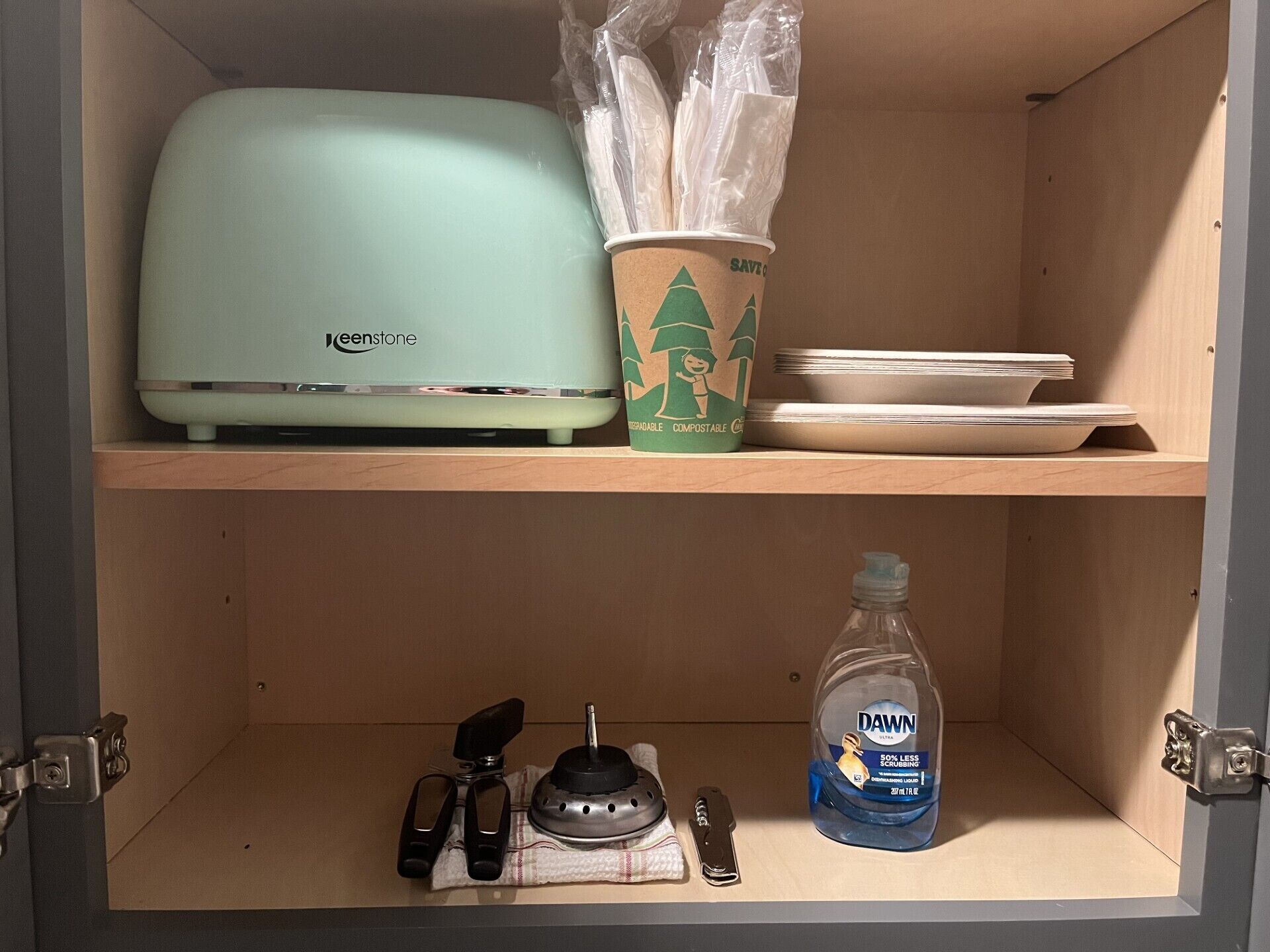 A kitchen cabinet with a Keenstone toaster, a coffee cup labeled 'SAVE CO', plates, Dawn dish soap, and a cloth.