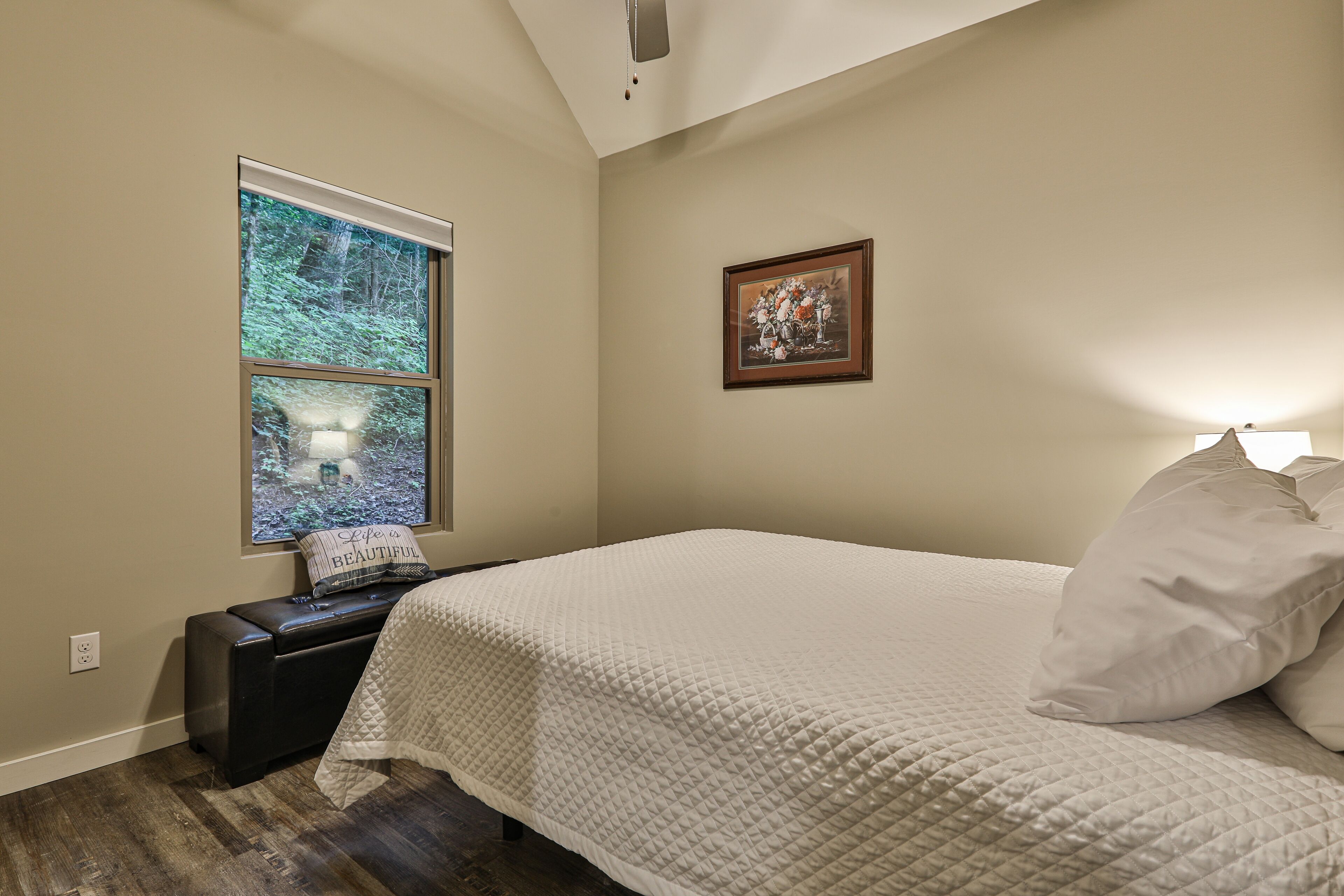 A bedroom with a bed, a window with a view of trees, a framed picture on the wall, and a small table with a pillow.