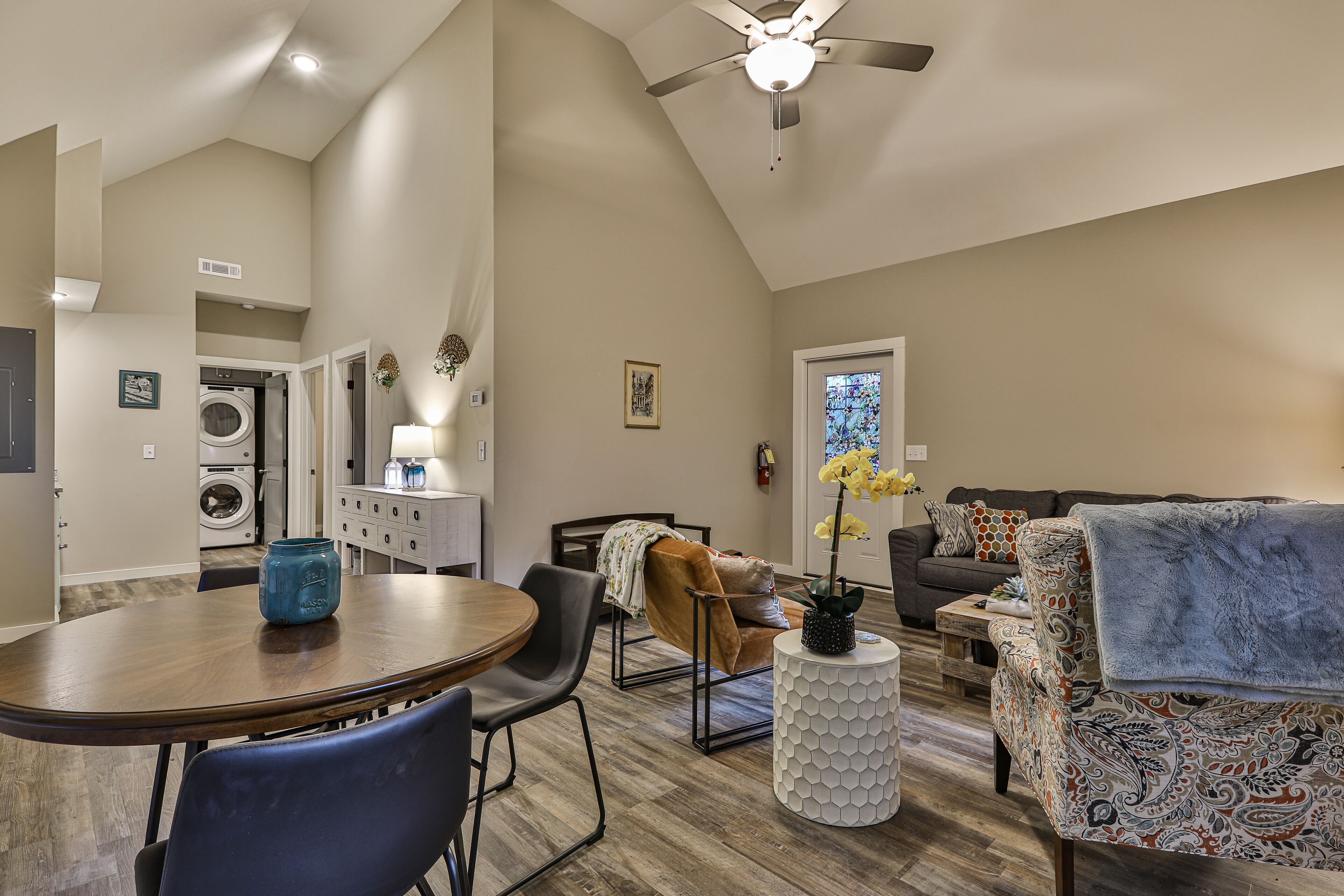A spacious living area with a dining table, chairs, a sofa, and a patterned armchair. A ceiling fan is visible, and a washing machine is seen in the background.