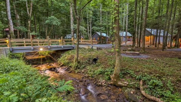 Un área boscosa con un puente de madera sobre un arroyo, rodeada de árboles y un pequeño edificio.