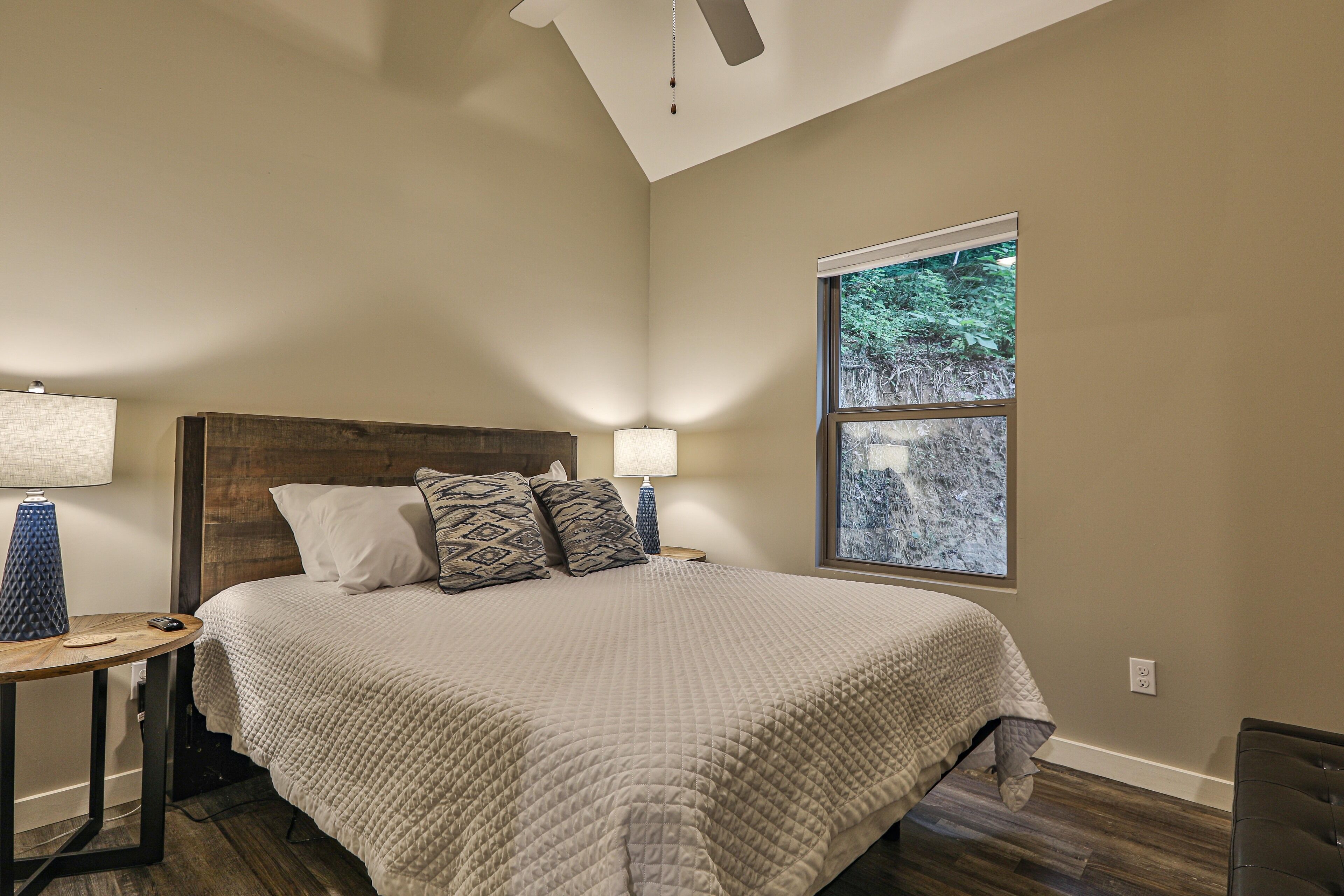 A bedroom with a large bed, two bedside lamps, a window with a view of trees, and a ceiling fan.