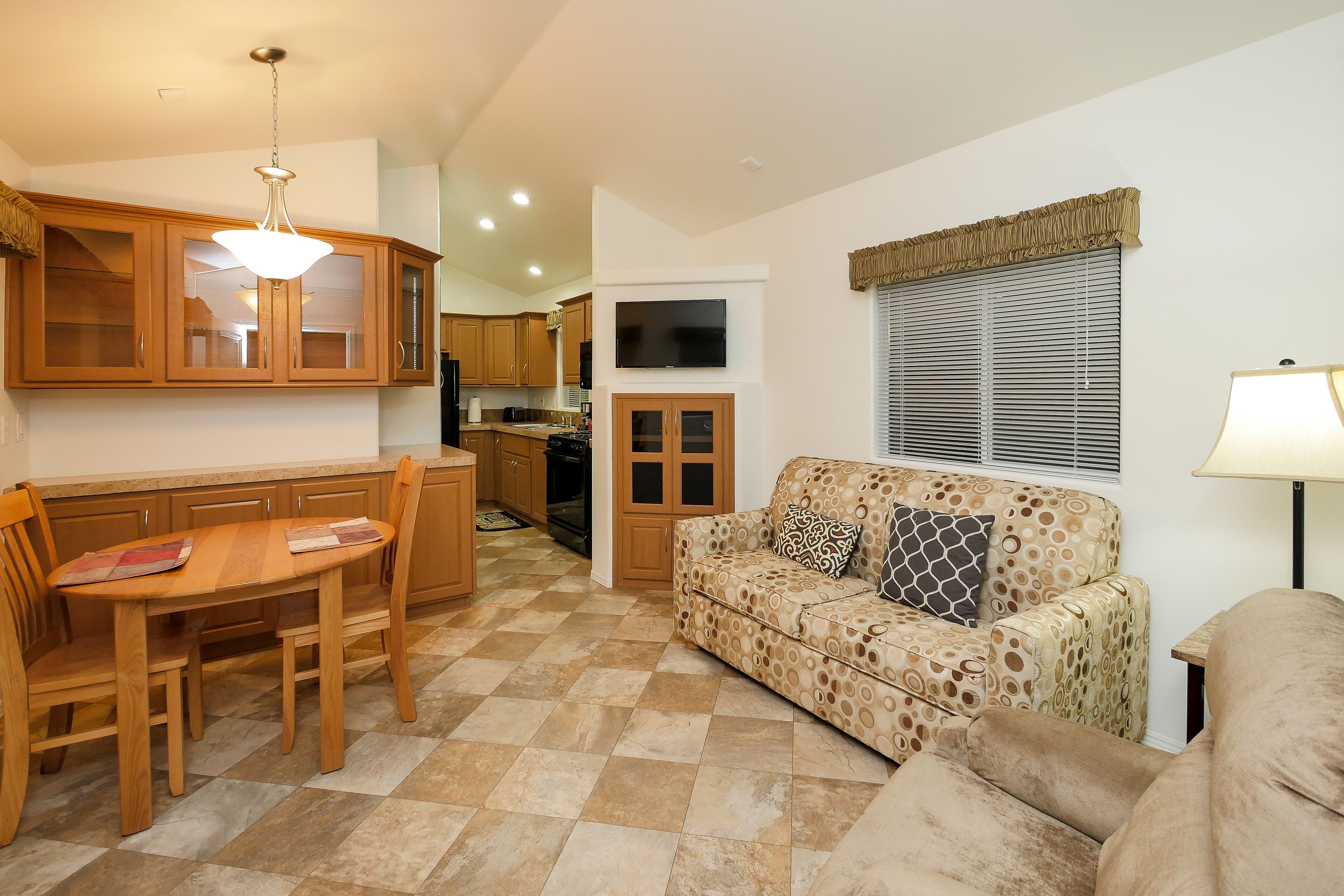 A living room with a patterned sofa, a dining table with chairs, wooden cabinets, and a TV mounted on the wall.