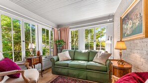 Ein gemütlicher Raum mit einem grünen Sofa, einem gemusterten Teppich und Blick auf Bäume durch große Fenster.