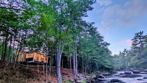 A cabin with a stone exterior and large windows, surrounded by dense forest and a rocky riverbank. - Ultra Private, Crooked Reflections, Riverside Retreat, Hot Tub, Waterfront Glass (Norway)