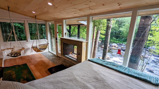 A bedroom with a large bed, a fireplace, a hanging chair, and a view of a forest and stream. - Ultra Private, Crooked Reflections, Riverside Retreat, Hot Tub, Waterfront Glass (Norway)