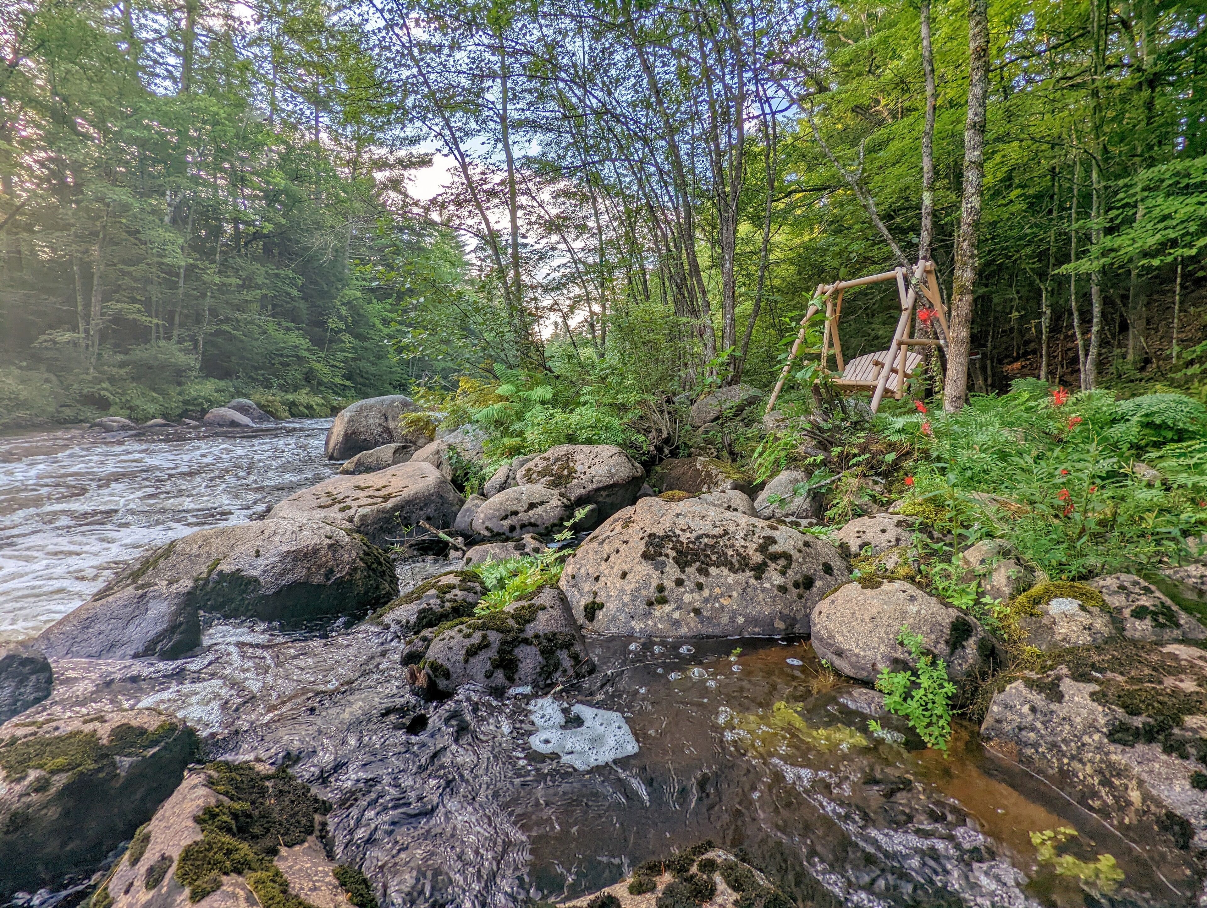 Una orilla rocosa de río con un conjunto de columpios de madera y un bosque denso al fondo.