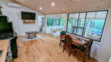Une pièce au plancher de bois, avec une table et des chaises, un téléviseur fixé au mur et une grande fenêtre donnant sur un paysage verdoyant.