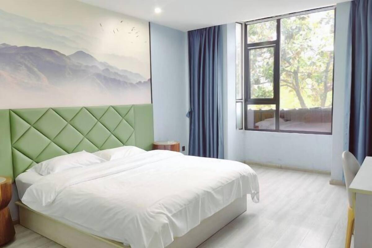 Un dormitorio con cama, escritorio, ventana con cortinas y un cuadro de un paisaje montañoso.