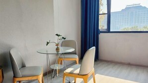 Una habitación con una mesa redonda de vidrio, dos sillas, una planta en maceta y vistas de un edificio a través de una ventana con cortinas azules.