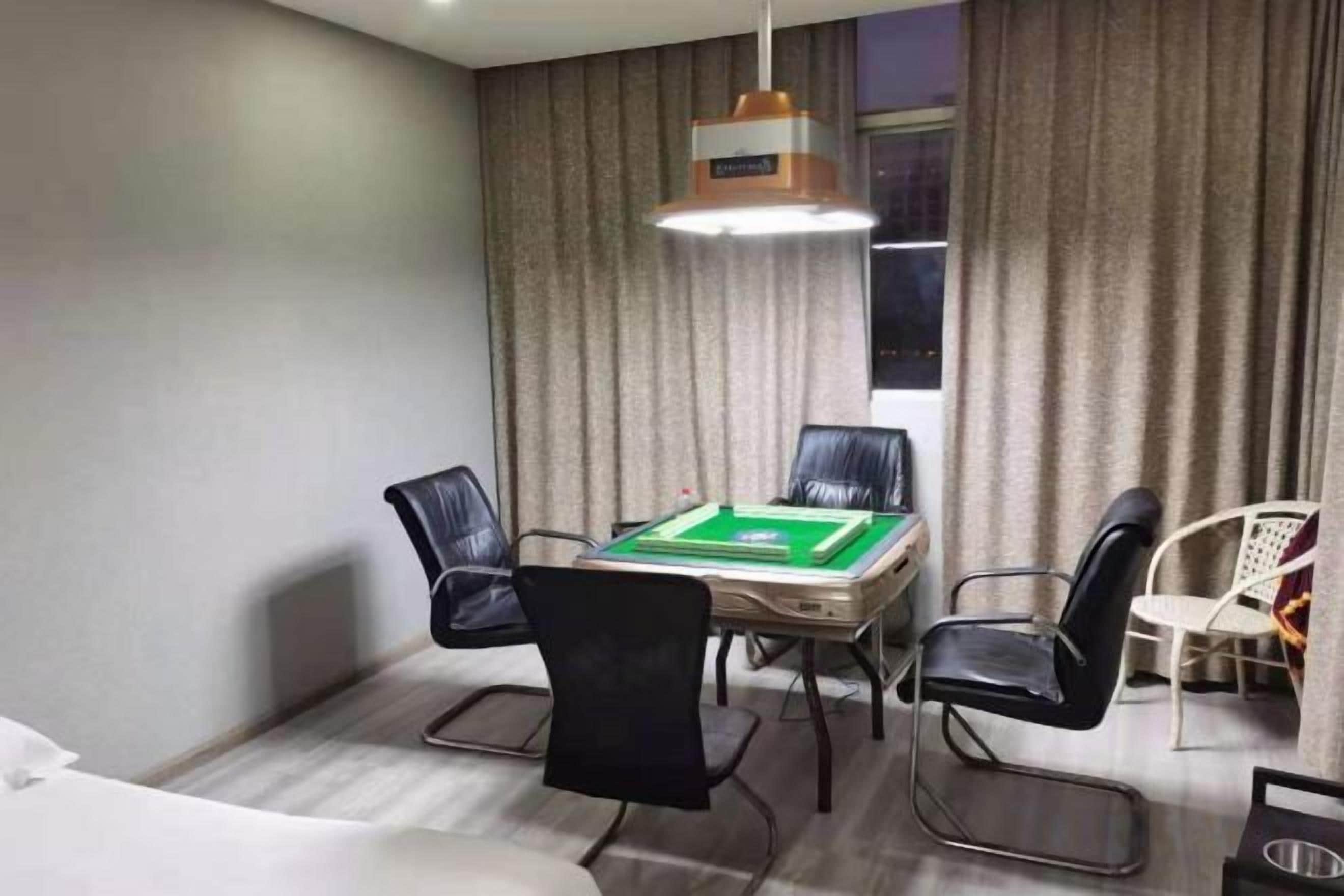 A hotel room with a green pool table, four black chairs, a white sofa, a hanging lamp, and a window with curtains.