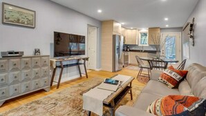 A modern living room with a sofa, coffee table, and TV. There is a dining area with a table and chairs. The kitchen is visible with a refrigerator and cabinets.