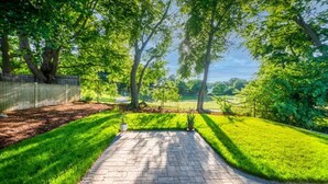 A paved pathway leading through a lush green garden with trees and a fence on one side.