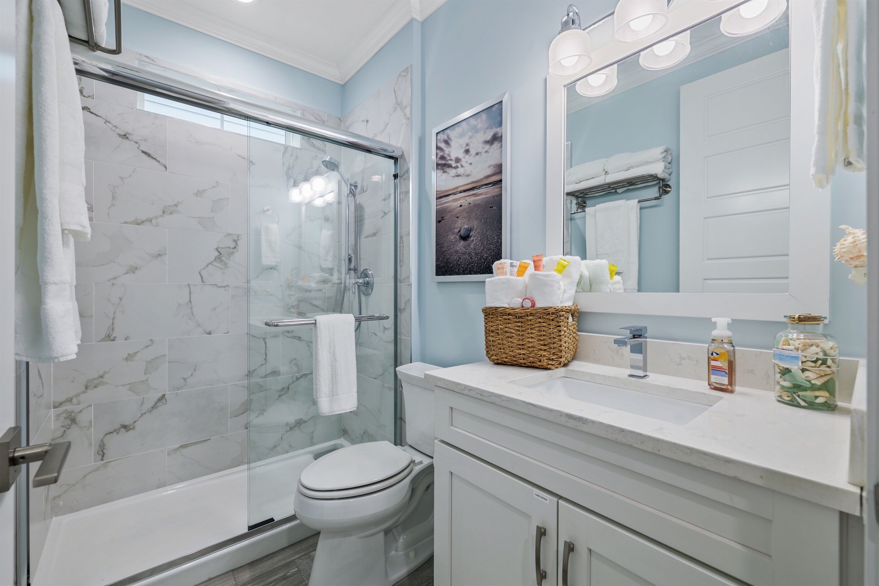 Une salle de bain avec une douche à parois de verre, des toilettes, un lavabo avec un miroir et un panier contenant des articles de toilette.