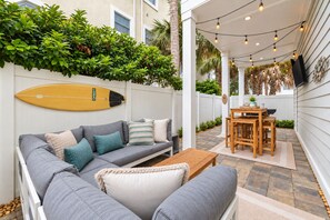 A cozy outdoor patio with a gray sofa, a wooden table, and string lights. - Ocean View Bliss | Steps to Beach | Golf Cart Perk (Jacksonville Beach)