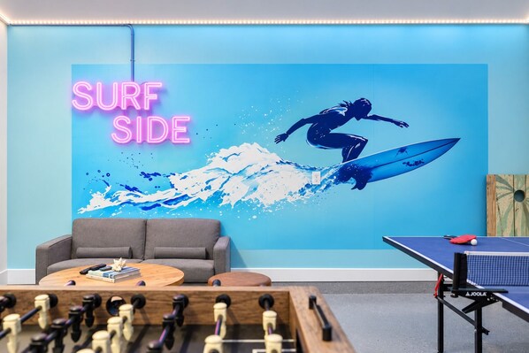 A room with a foosball table, a ping pong table, a sofa, and a wall with a 'SURF SIDE' neon sign and a surfer illustration. - Ocean View Bliss | Steps to Beach | Golf Cart Perk (Jacksonville Beach)