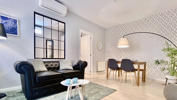 A modern living room with a black sofa, a dining table with chairs, a patterned wall, and a large window. - Lavapies - One-bedroom Apartment (MADRID)