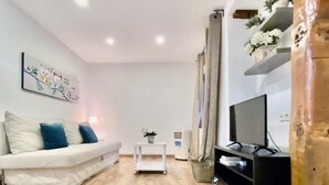 Ein Wohnzimmer mit einem weißen Sofa, einem kleinen Tisch mit Lampe, einem Fernseher auf einem Beistelltisch und einer Zimmerpflanze.