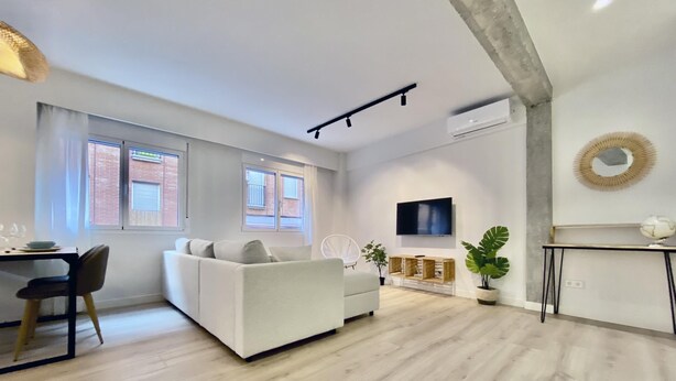 A modern living room with a sofa, a dining table with chairs, a TV mounted on the wall, and large windows.