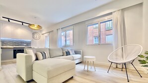 A modern living room with a sofa, a chair, and a small table. There is a kitchen area with a clock and a window.