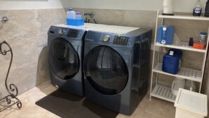 Un área de lavandería con dos lavadoras de carga frontal, un estante con productos de limpieza y una pared de azulejos.