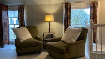 A room with two green armchairs, a small table with a lamp, and a view of a building through a window with patterned curtains.