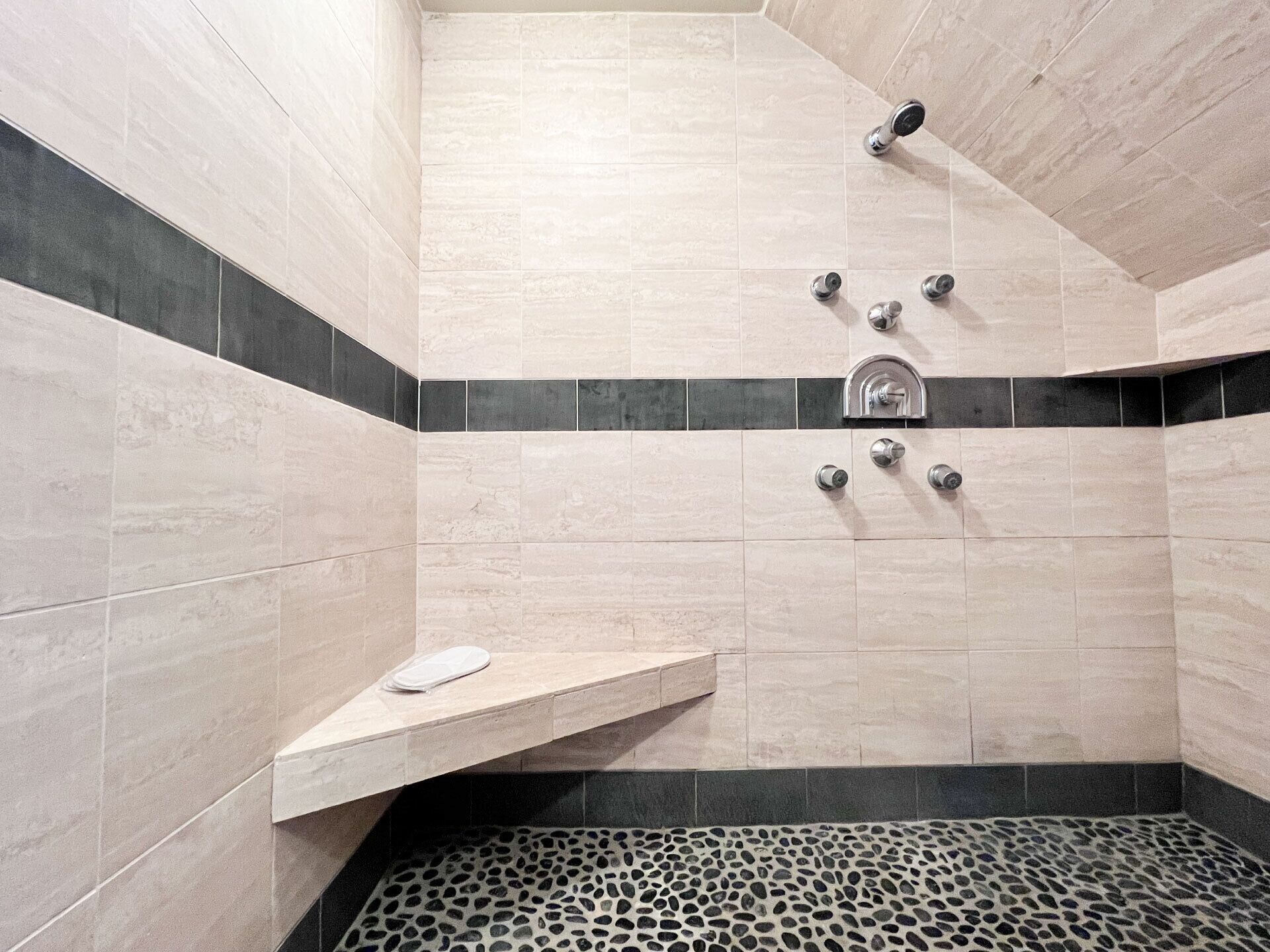 A modern bathroom with beige tiles, a black and beige border, a shower with multiple nozzles, and a pebble-patterned floor.