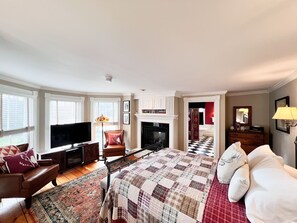 A spacious bedroom with a large bed, a television, a fireplace, and a view of the bathroom. - Four Columns Inn (Newfane)