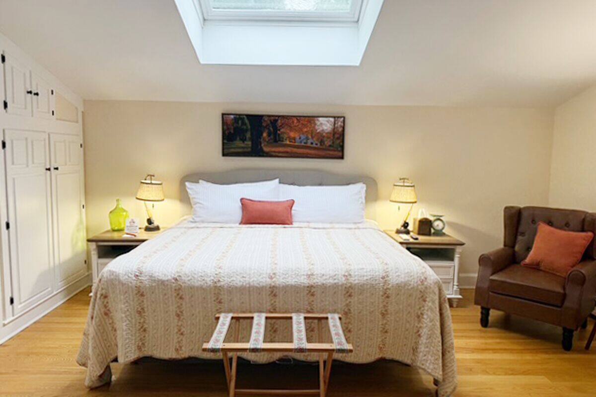 Un dormitorio con tragaluz, una cama con colcha estampada, dos mesitas de noche con lámparas, una silla y un banco de madera.