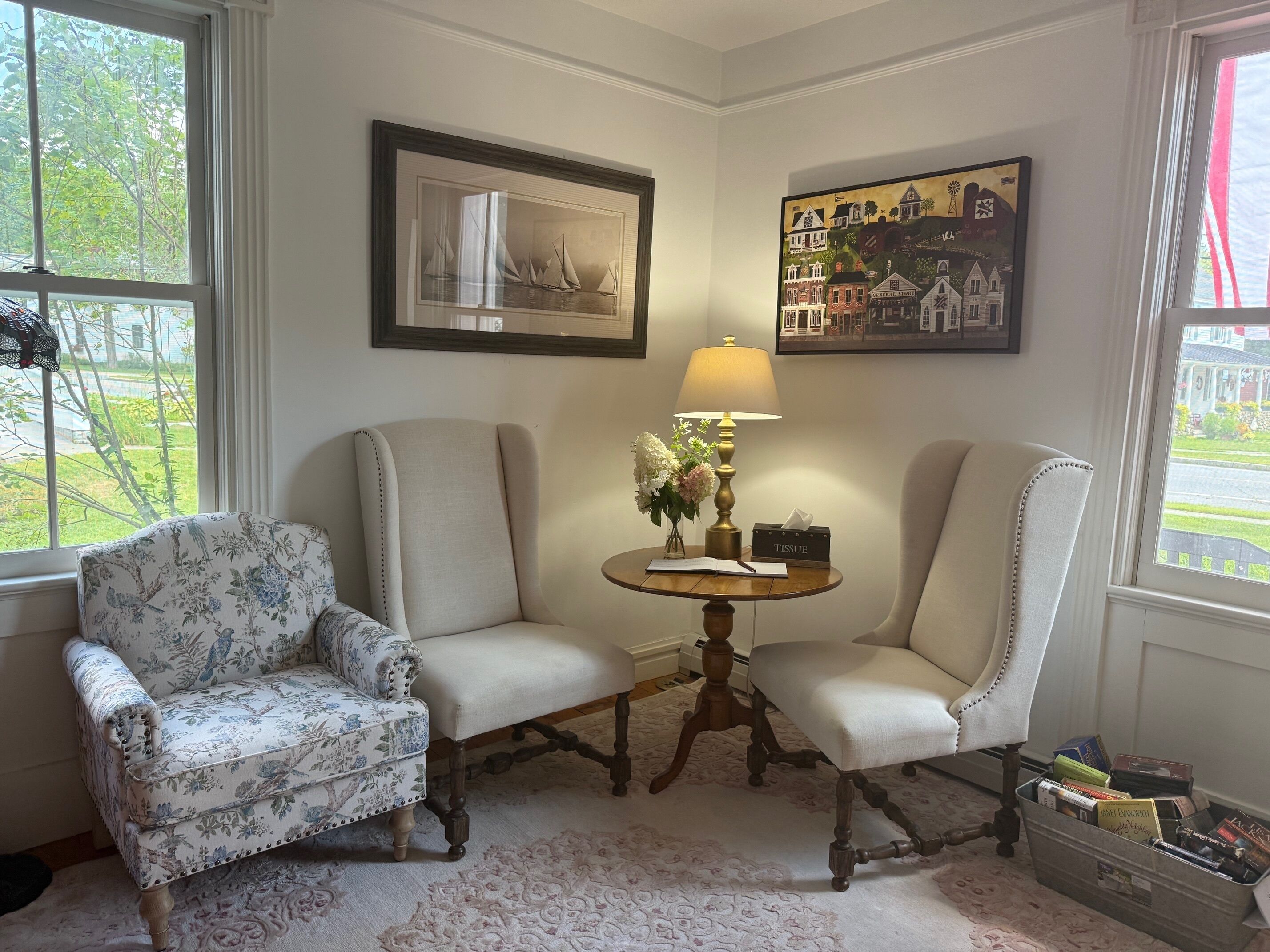 A room with two upholstered chairs, a small round table with a lamp, a framed picture, and a window with a view of greenery.