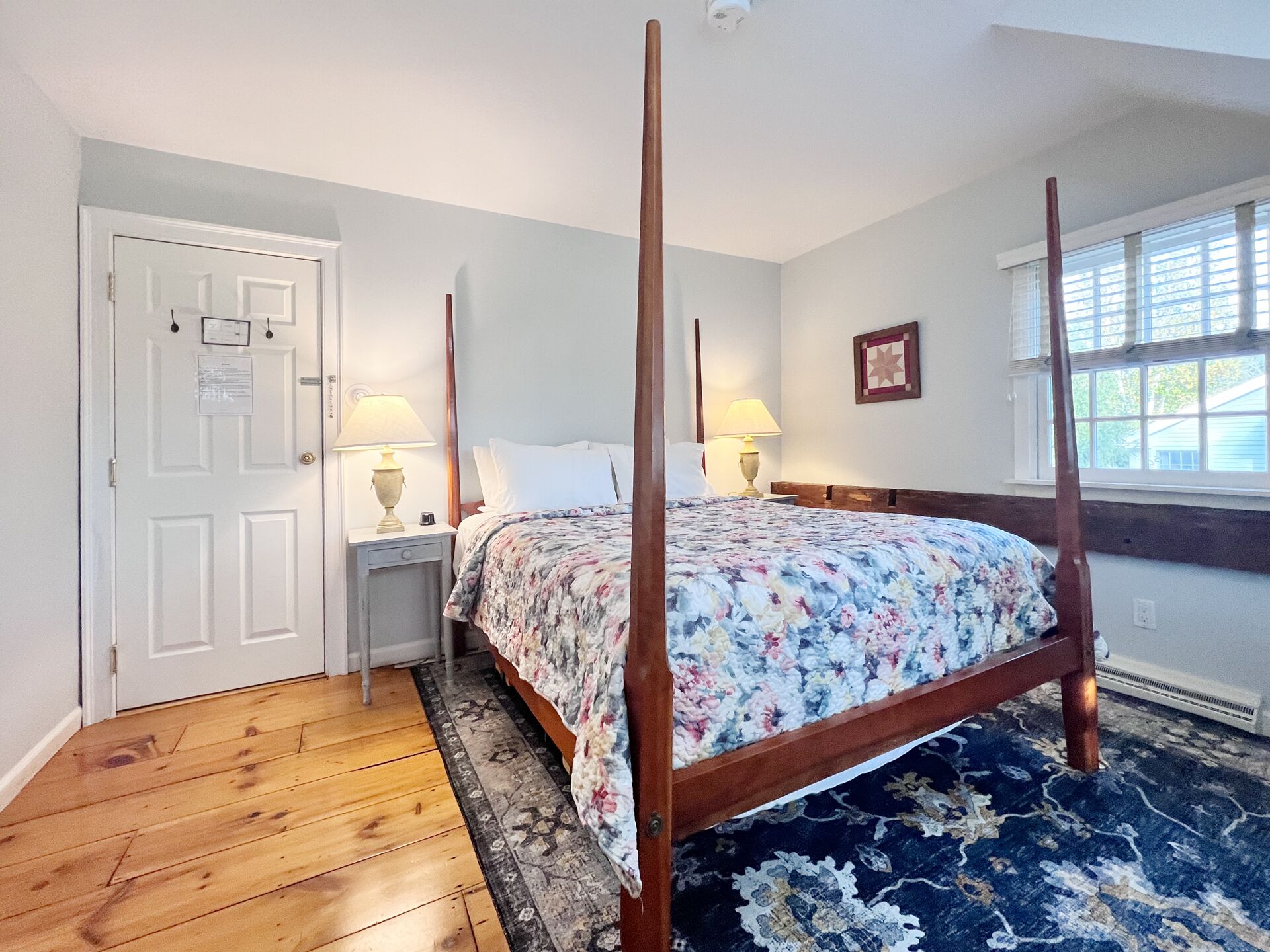 A bedroom with a four-poster bed, a wooden floor, a window with blinds, and a door with a notice.