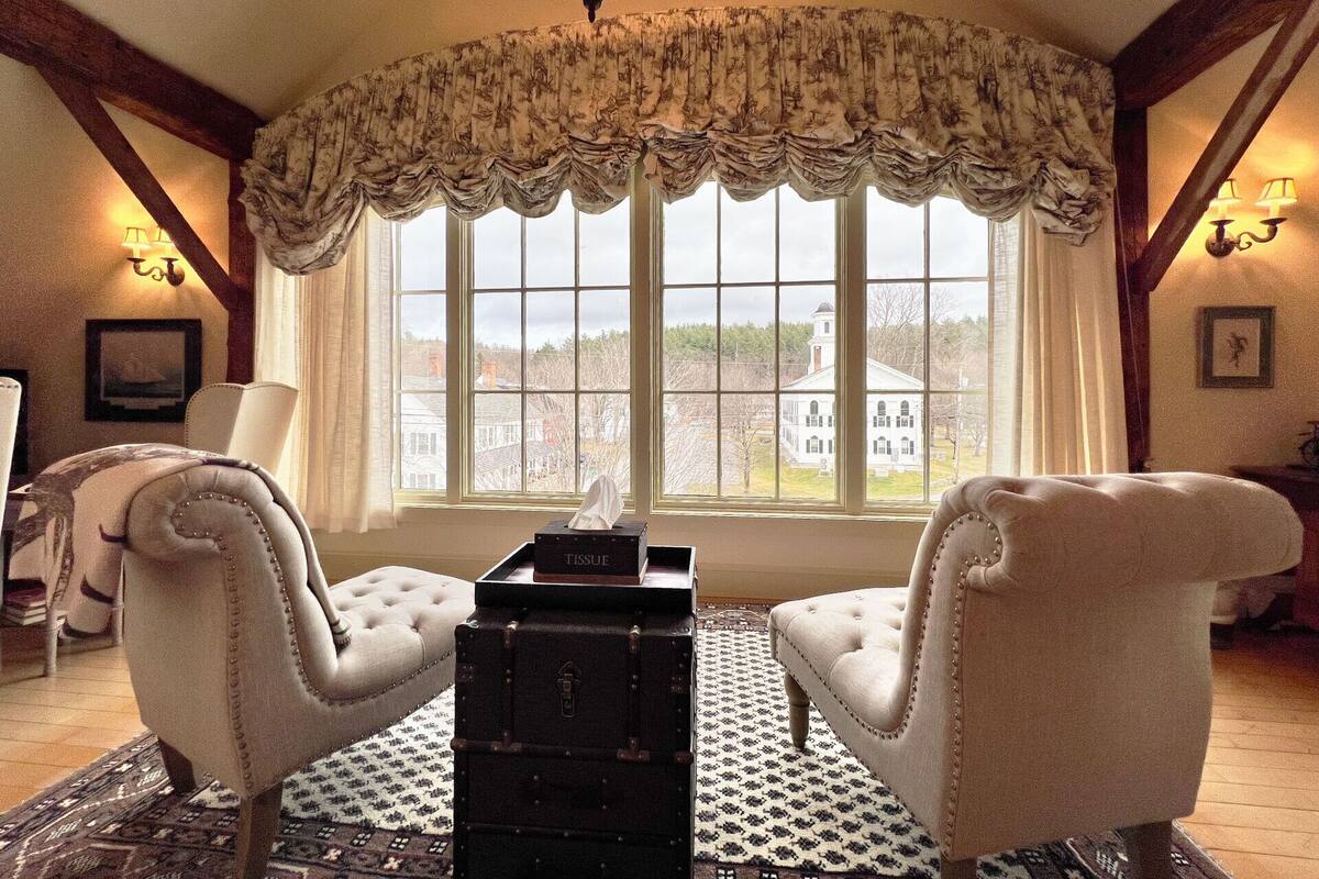 Una habitación con dos sillones tapizados, un baúl negro y un ventanal con vistas a un edificio blanco.
