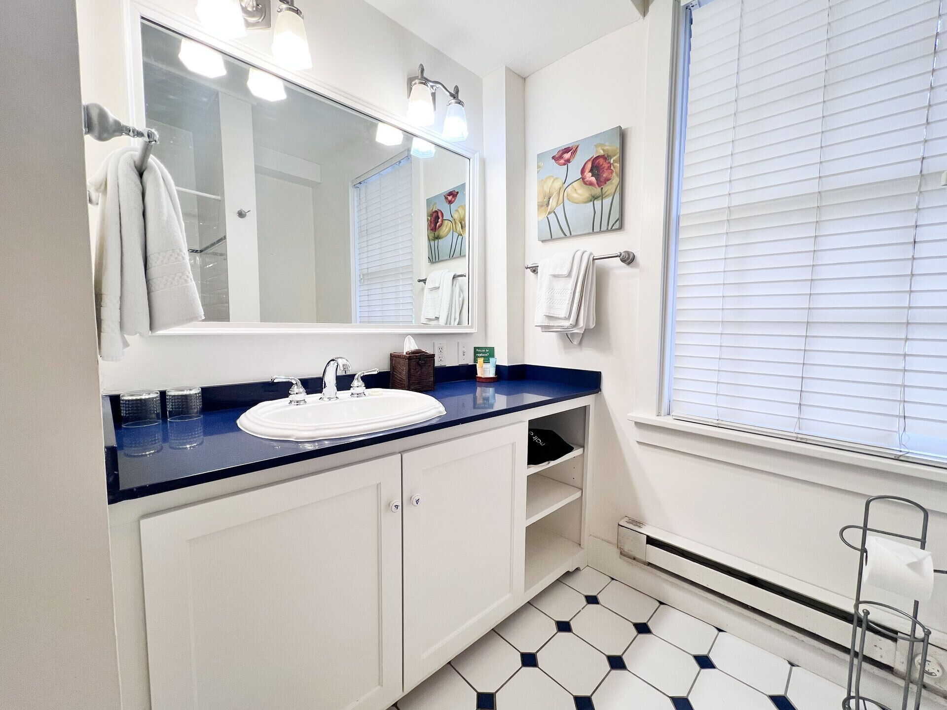 A bathroom with a white sink, blue countertop, and a large mirror. There are towels hanging on a rack, a painting on the wall, and a window with blinds.