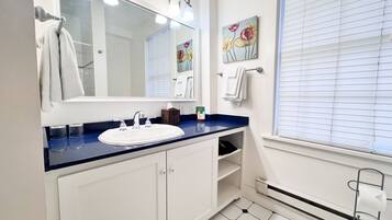 A bathroom with a white sink, blue countertop, and a large mirror. There are towels hanging on a rack, a painting on the wall, and a window with blinds.