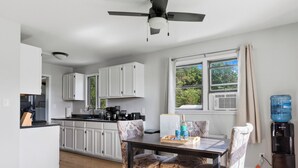 Une cuisine moderne avec des meubles blancs, une table à manger entourée de chaises à motifs, un ventilateur de plafond et une grande fenêtre avec des rideaux.