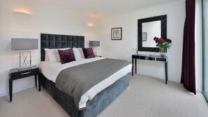 Une chambre à coucher moderne avec un grand lit, deux tables de chevet avec des lampes, une console sur laquelle est posé un vase de fleurs et un grand miroir au mur.
