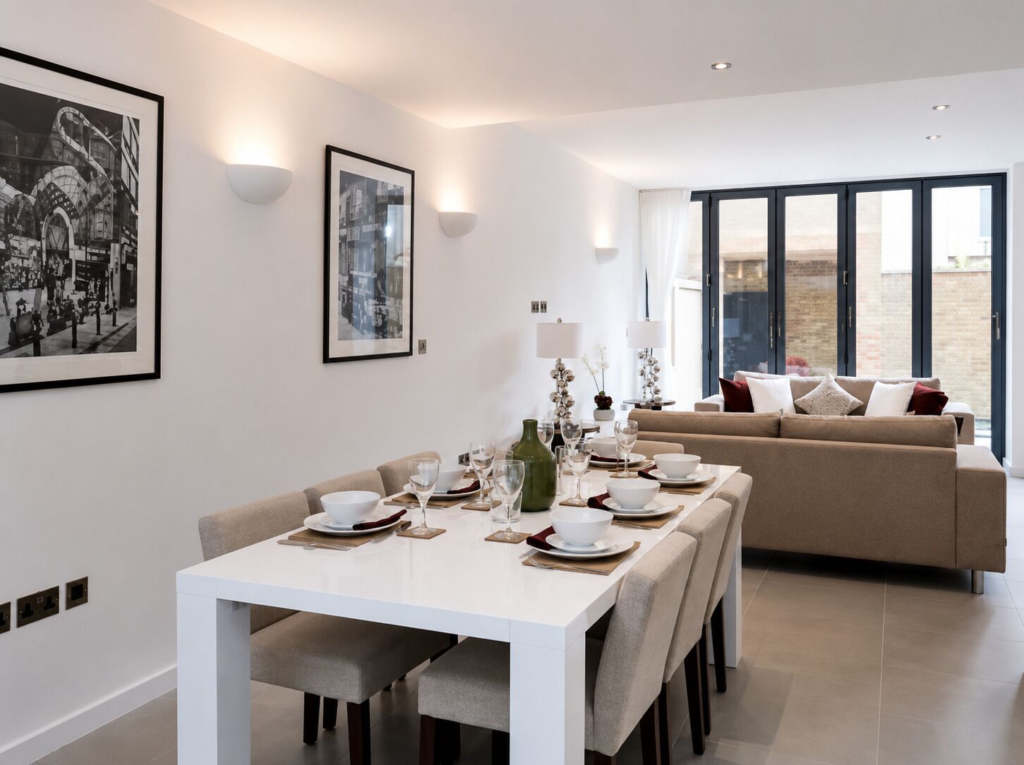 Un'elegante sala da pranzo con un tavolo bianco apparecchiato per quattro persone, un divano sullo sfondo e due quadri appesi al muro.