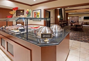 A buffet area with a sign indicating free daily breakfast, a glass display case, and a dining area with tables and chairs.