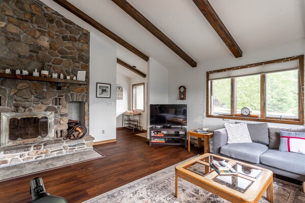 A living room with a stone fireplace, wooden beams, a television, a sofa, and large windows.