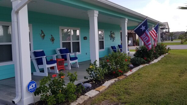 Una casa turquesa con un porche, dos sillas azules y un pequeño jardín con flores rojas.