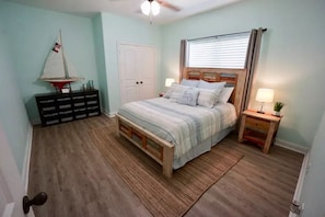 Quarto com cama, mesa de cabeceira, cômoda, decoração náutica com um veleiro e uma porta.