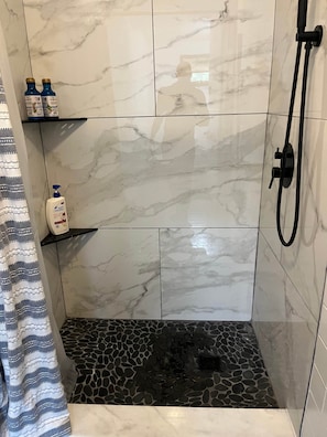 A shower with a black pebble floor, white marble tiles, and a handheld showerhead.