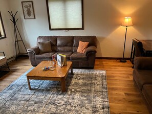 A living room with a brown leather sofa, a wooden coffee table with books and glasses, a patterned rug, and a floor lamp.