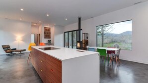 A modern kitchen with a central island, a dining area with colorful chairs, and a large window offering a view of the outdoors. - Asheville Meadows Pool, Hot Tub & Forest Views (Asheville)