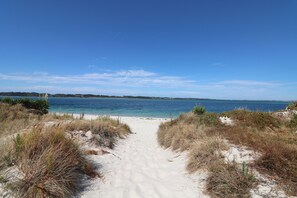 Ein Strandweg führt zu einem Sandstrand mit klarem, blauem Wasser und sanften Hügeln in der Ferne.