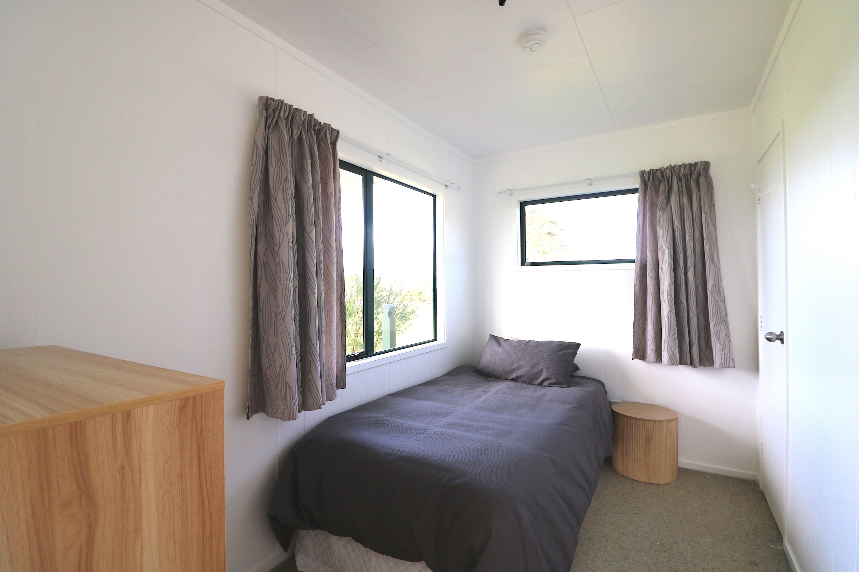A bedroom with a bed, two windows, a bedside table, and a small round wooden stool.