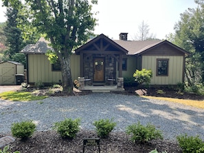 A two-story house with a stone chimney, a front porch with a wooden door, and a small garden area with gravel and shrubs.