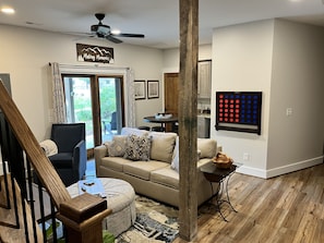 A living room with a sofa, ottoman, and a dining table with chairs. There is a staircase with wooden railings, a ceiling fan, and a view of the outdoors through a glass door.